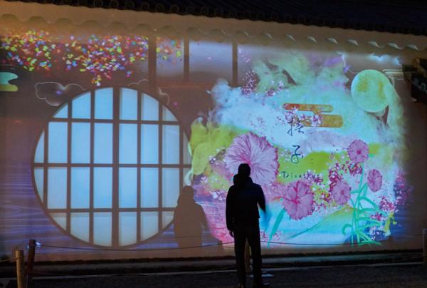 写真：重要文化財 台所（屋外）にて、塀に映し出された円窓の映像の前に立つ来場客。
