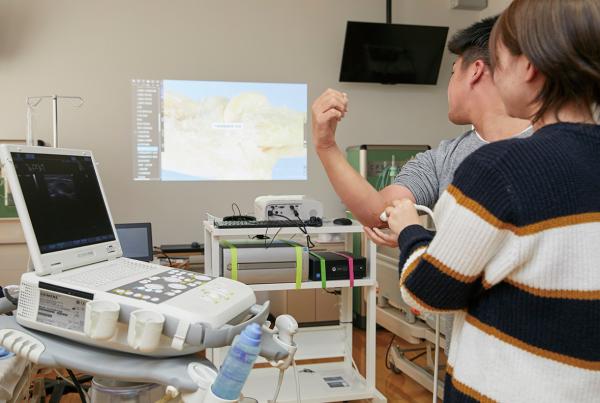 写真：女性が男性の肘の内側に超音波エコー機を当て、壁に映し出された解剖映像を確認している様子。