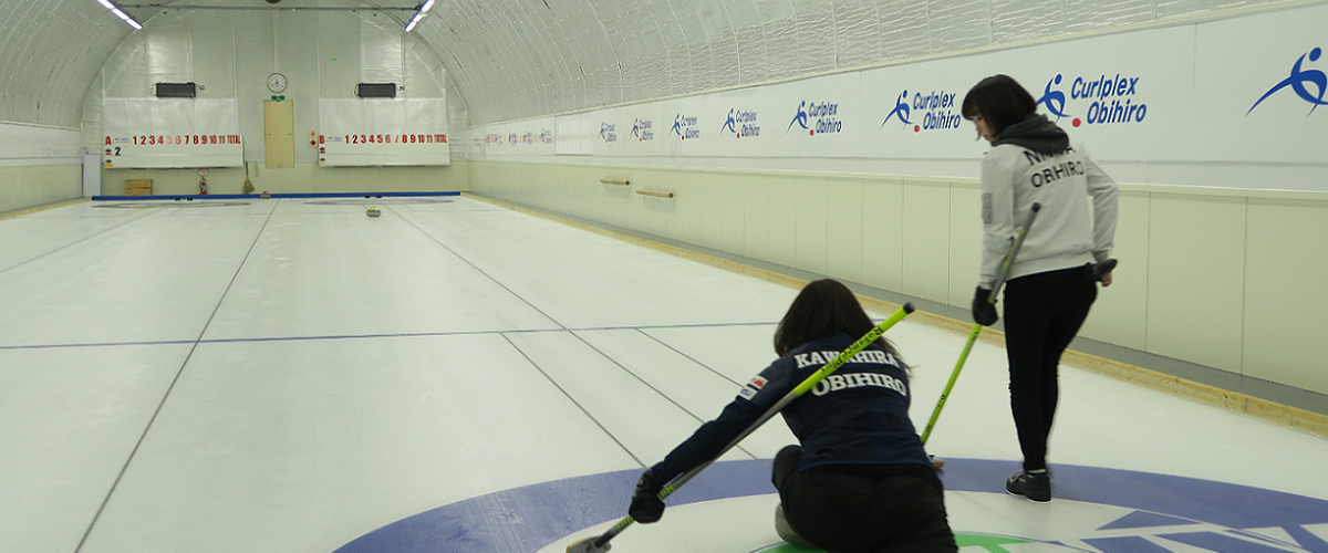 写真：二人の女性カーリング選手が競技場でカーリングをする様子。左の選手がストーンを持ち投げようとしており、右の選手はその右横に立ちストーンを見ている。