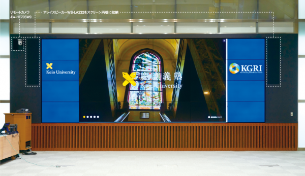 写真：写真：55V型・縦４面、横６面の24面の大型マルチスクリーンシステムの左右縦３面は青背景、左に「Keio university」、右に「KGRI」の文字が映し出されている。中央16面には教会のステンドグラスの背景、慶応義塾大学のロゴとともに「慶應義塾」という文字が映し出されている。済むリーンの両端にはアレイスピーカーWS-LA232が収納されている。また、向かって左側上部にはリモートカメラAW-HE70SW9が設置されている。