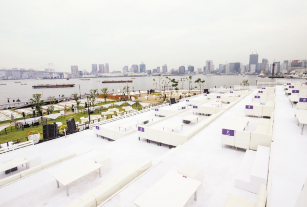 写真：東京湾前の会場。遠くにビル群があり、手前に海、芝生の陸、段差をつけた観客席には白いシートと机が置かれている広いVIPエリアがある。