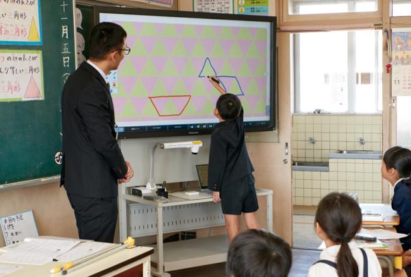 写真：算数の授業の様子。沢山の色違い三角形が画面一杯に整列して映し出されたタッチスクリーンディスプレイ。映し出された三角形をなぞって台形や三角形をディスプレイに描く男児。
