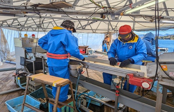 青い作業服を着た2人が屋外の作業場で機械を操作している。