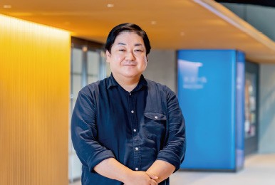 明るい室内で微笑む男性。背景に青い看板と木材の壁。