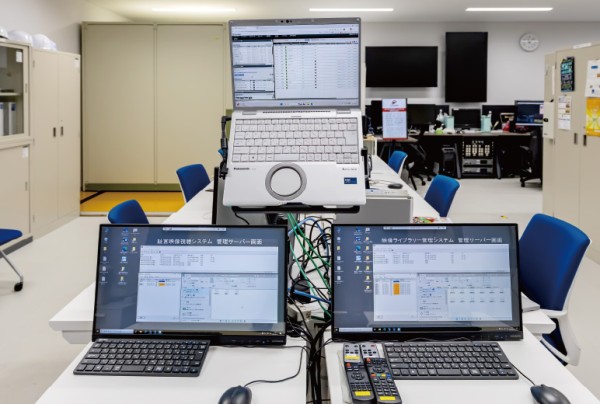 オフィスの机に設置された3台のモニターとキーボード、背景には椅子や収納棚がある部屋。