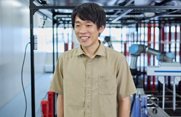 男性が研究室内で笑顔。背景にロボットアームがある先進的な設備。