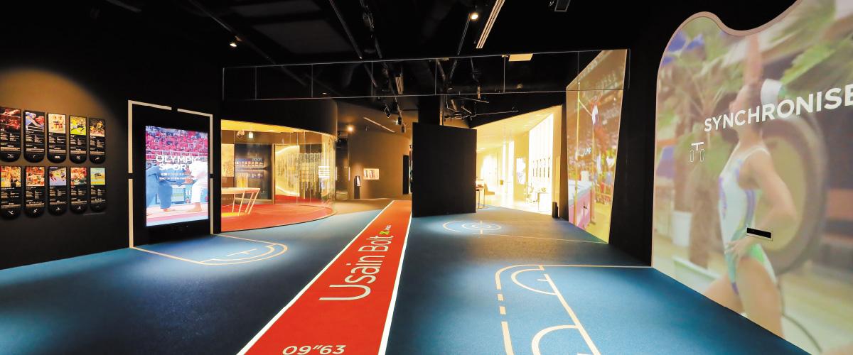 写真：オリンピックミュージアム展示ブースの一部、左に柔道関連の展示、右側に陸上競技に関するブースがあり、それぞれのブースで映像が映し出されている。中央の床にはウサイン・ボルトが実際に出した記録と距離が